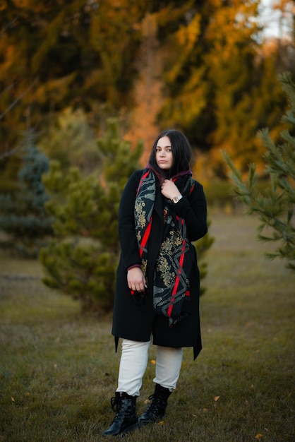 A darkhaired girl in a black coat and white pants is holding a scarf and posing in the park