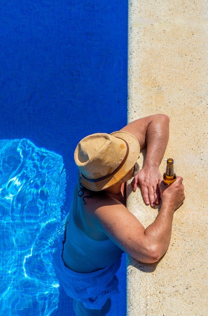 Darkhaired man with straw hat and sunglasses half body length standing in crystal clear blue water leaning on the edge of a luxury swimming pool relaxing and holding a bottle of beer