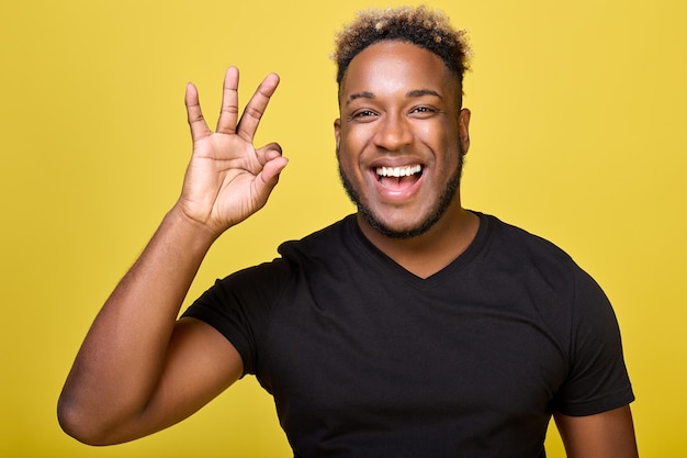 Темнокожий улыбающийся мужчина показывает пальцами знак «ОК». Спортивное телосложение, симпатичный афроамериканец на желтом фоне указывает жестами рук на успешную идею.