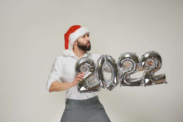 A dashing man in a red velvet santa hat with his lips in a tube\
is pushing silver balloons in the shape of 2022 to the right. a guy\
with a beard at a new year party.