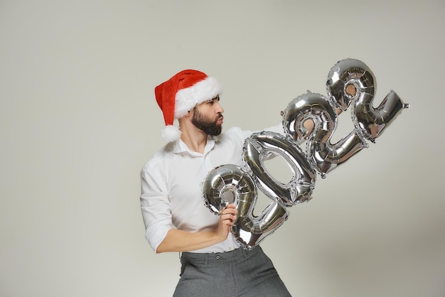 A dashing man in a red velvet santa hat with his lips in a tube\
is pushing silver balloons in the shape of 2022 to the vertical\
right. a guy with a beard at a new year party.