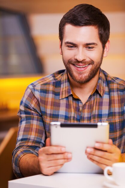 Dat is leuk. Knappe jonge man die naar zijn digitale tablet kijkt en glimlacht terwijl hij in de coffeeshop zit