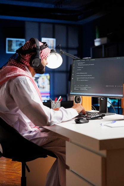 Data developer from the Middle East types code on a computer demonstrating the core of software programming and development Muslim man using wireless headphones and immersed in algorithm execution