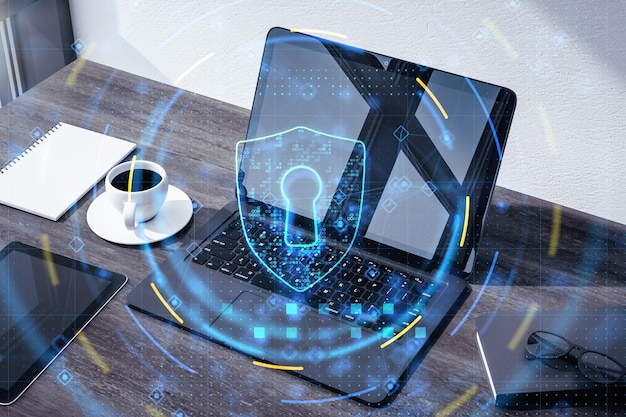 Data safety and cyber security concept with digital graphic glowing blue shield symbol with keyhole on office table with laptop and coffee mug background double exposure