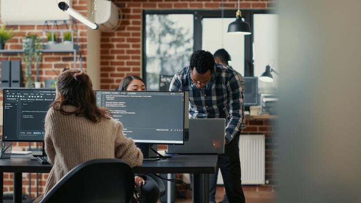 Photo database programer writing code in front of multiple computer screens displaying artificial intelligence algorithm. developer coding database while colleagues doing teamwork in background.