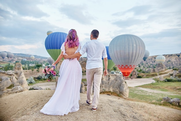 Date of a couple in love at sunset against background of balloons in Cappadocia, Turkey. Man and woman hugging standing on hill and look at big balloons. Engagement in the mountains of Cappadocia