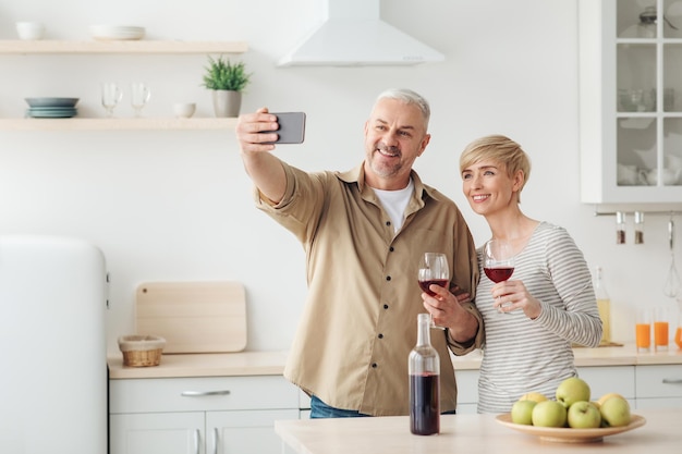집에서 데이트하고, 축하하고, 셀카를 찍고, 가족을 위한 편안한 아파트입니다. 행복한 성인 남편과 아내는 레드 와인이 든 안경을 들고 현대적인 주방 인테리어, 여유 공간에서 소셜 네트워크를 위해 사진을 찍습니다.