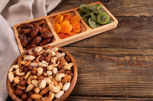 Dates, dried apricots and kiwis in a Compartmental dish and assortment of nuts in wooden bowl on a dark wooden table.