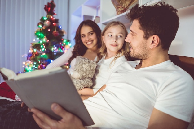 The daughter and parents lay on the bed and read a book near the christmas tree