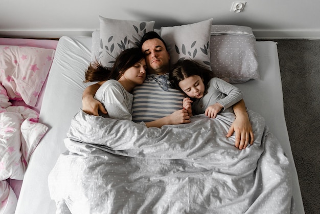 ベッドで両親と娘、お父さんは彼の妻と娘に優しくキスします、強い家族の抱擁、ベッドで家族の週末、眠っている家族、娘はベッドで若い両親と一緒に寝ます