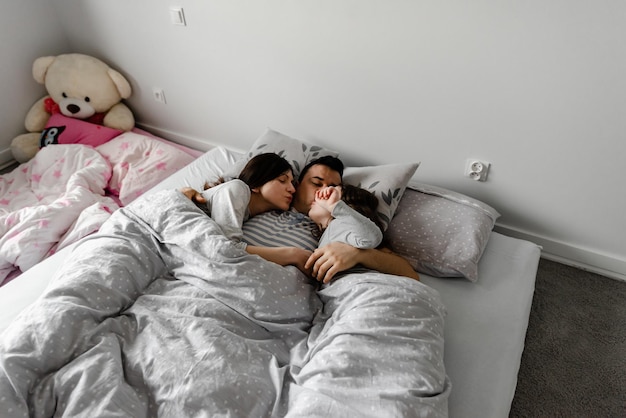 Daughter with parents in bed,dad gently kisses his wife and daughter,strong family hugs,family weekend in bed,sleeping family,daughter sleeps with young parents in bed