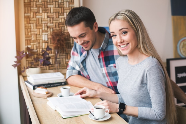 Day in cafe. Young couple in cafe with stylish interior. Students having delicious coffee drinks. They reading book. Woman smiling and looking at camera