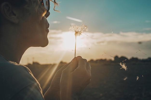 Photo day dreaming leisure activity with woman blowing a dandelion outdoor in the nature park emotion and love lifestyle people concept freedom and travel dreams sky and sunset in background