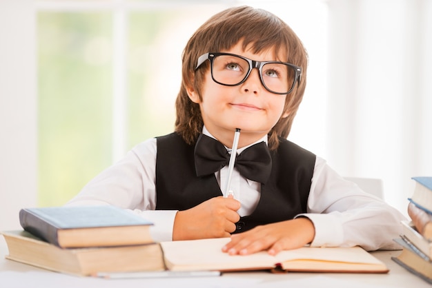 Day dreaming. Little boy leaning his face on pen and looking away while sitting at the desk