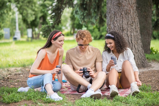 Day off, picnic. Three young joyful friends with drink looking photos on camera sitting on blanket outdoors on warm day