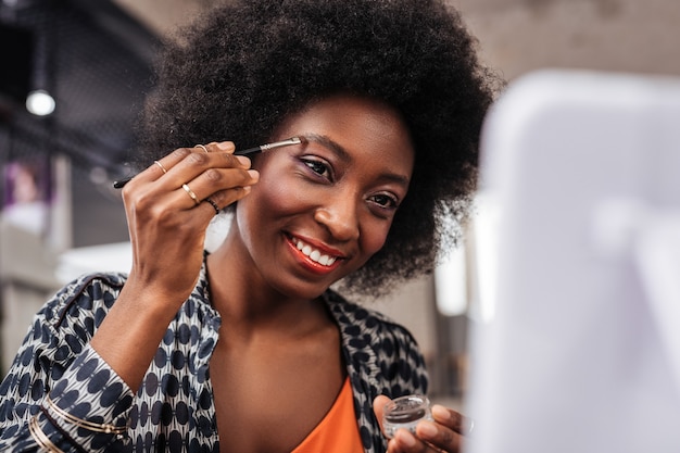 Day in a studio. Smiling dark-skinned woman in an orange top looking involved while trying new eyebrows shape