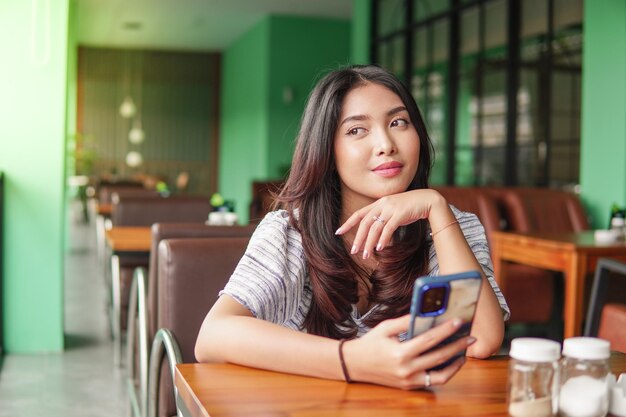 Молодая азиатка в платье сидит в ресторане на завтрак с смартфоном в руках, поддерживая подбородок, потерянная в мыслях.