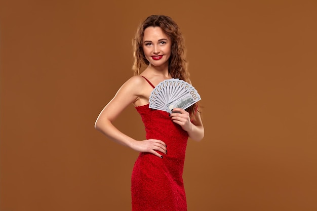 Dazzling curly woman weared in a red sexy dress is holding some hundred dollar bills in her hands, put her hand on the waist, smiling and looking at the camera. Casino concept on a brown background.