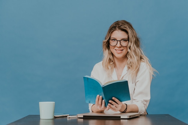 De aantrekkelijke blondevrouw in glazen die witte overhemdszitting dragen op haar werkende plaats met laptop, houdt haar agenda, thuis werkend tijdens quarantaine over blauwe achtergrond. Coronavirus pandemisch concept.