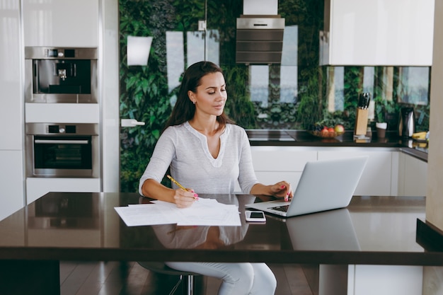 De aantrekkelijke jonge moderne zakenvrouw die thuis met documenten en laptop in de keuken werkt