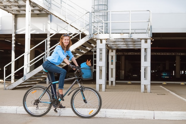 De aantrekkelijke moderne vrouw met een kleine hond in een mand die op de fiets door de stad beweegt