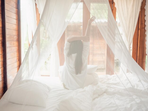 Foto de achterkant van de vrouw die zich uitstrekt in het bed na het ontwaken begroet de nieuwe dag met warm zonlicht.