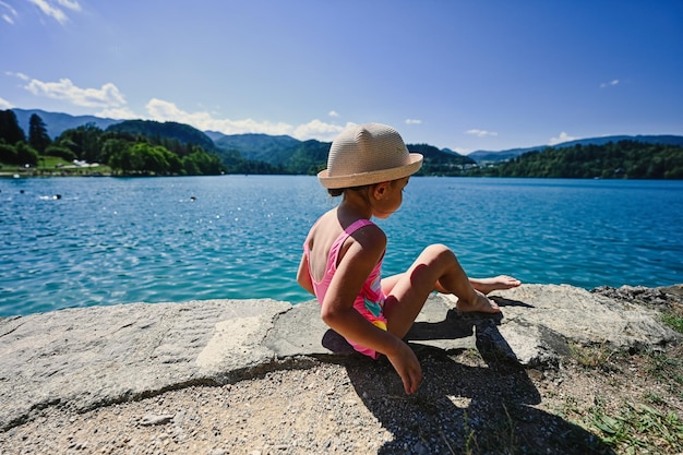 De achterkant van het babymeisje is een badpak en een hoed zit op de pier van het prachtige Bled-meer in Slovenië