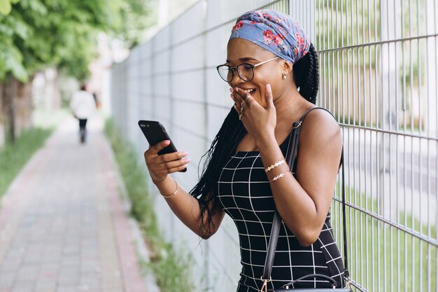 De Afrikaanse Amerikaanse vrouw in een kleding heeft pret buiten blik in telefoon en glimlach