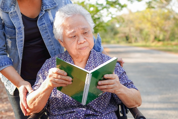 De Aziatische oudste of bejaarde oude patiënt die van de damevrouw een boek lezen terwijl het zitten op rolstoel in park: gezond sterk medisch concept