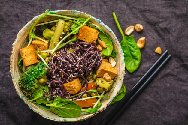 De Aziatische veganist beweegt gebraden gerecht met tofu, rijstnoedels en groenten, donkere achtergrond.