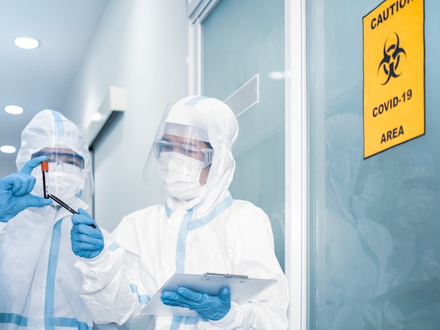 De Aziatische vrouw arts in persoonlijk beschermend kostuum met masker schrijft op de quarantainepatiëntgrafiek, houdt bloedmonster voor onderzoek coronavirus met covid-19 gebieds alert teken. Coronavirus, covid-19 concept.