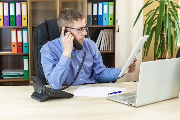 De baas op kantoor aan zijn bureau die aan de telefoon praat met een financieel rapport in zijn handen