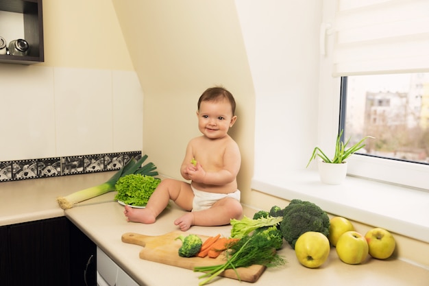 De baby in de luier zit op de keukentafel. Een kind speelt en heeft plezier met verse biologische groenten en fruit.