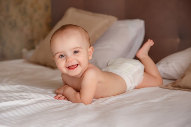 De baby in een luier ligt lachend op het bed in de slaapkamer op zijn buik