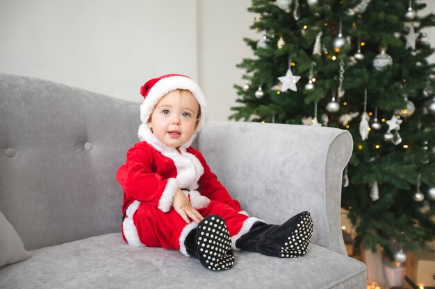 De baby in santakostuum zit op bank met Kerstboom