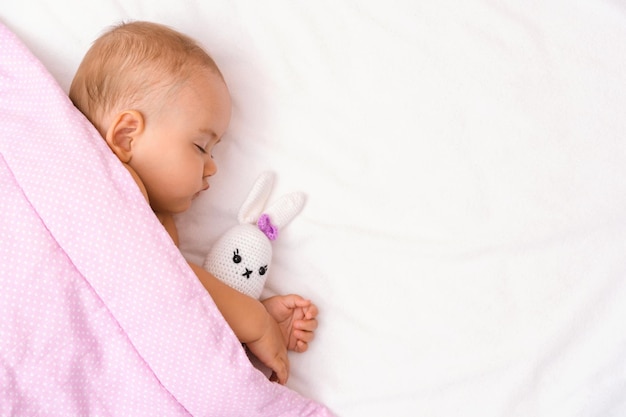 De baby slaapt op het bed met zijn favoriete speeltje onder de deken. Een baby in een droom knuffelt een gebreide haas, bovenaanzicht. Ruimte kopiëren