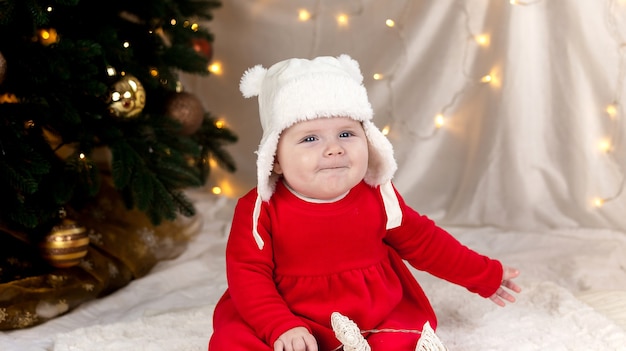 De baby van Kerstmis glimlacht. Een schattig klein meisje in een rode jurk en een witte hoed drukt emoties uit.