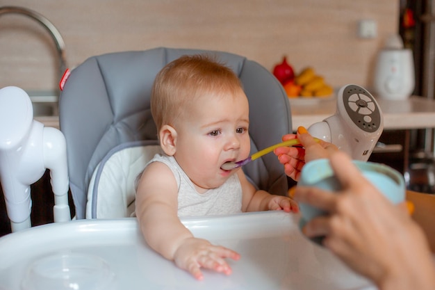 De baby zit in een hoge stoel in de keuken waar zijn moeder pap aan het voeren is