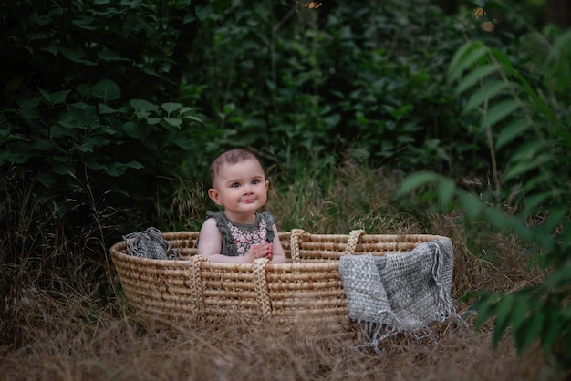 Foto de baby zit in een rieten wieg in een groen park. klein meisje in schattige jurk glimlachen. portret.