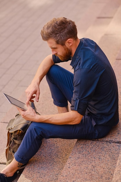 De bebaarde casual reiziger man zit op een trede en met behulp van een tablet-pc.