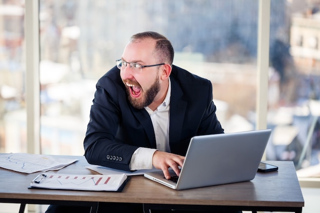 De bedrijfsgek zit achter de computer op kantoor en wordt gek, het emotionele portret van de man aan tafel. Gekke werker op zijn werkplek