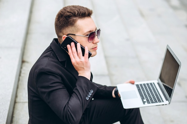 De bedrijfsmens zit op treden buiten bureau die aan laptop computer werken en op de telefoon spreken