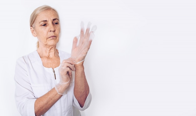 De bejaarde hogere rijpe vrouwenarts of de verpleegster in een witte medische laag zetten handschoenen aan die persoonlijk geïsoleerd beschermingsmiddel dragen. Gezondheidszorg en geneeskunde concept. Covid-19 pandemische crisis