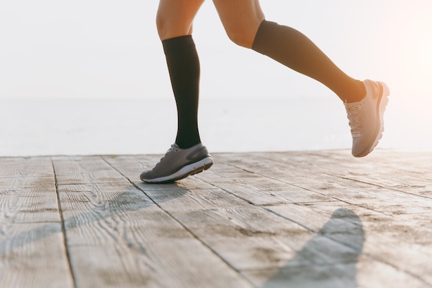 De benen van het meisje in zwarte beenkappen en grijze sneakers, die 's ochtends bij zonsopgang in de buurt van de zee lopen