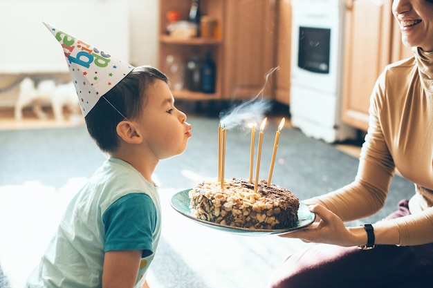 De blanke moeder houdt de taart vast terwijl de zoon op zijn verjaardag de kaarsjes blaast die een feestmuts dragen