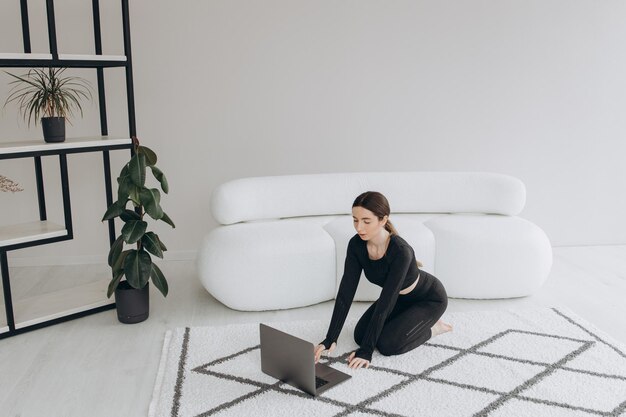 De blanke sportieve vrouw die fitnesskleding draagt, zit 's ochtends gehurkt met behulp van een laptop in de buurt van haar