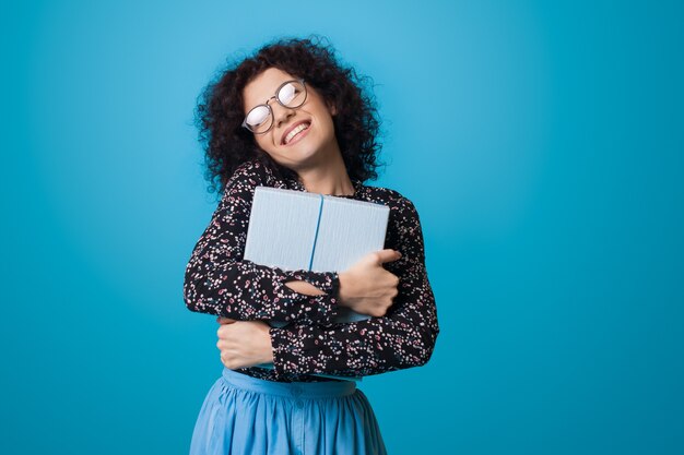 De blanke vrouw met krullend haar omhelst een cadeau dat glimlacht vooraan op een blauwe muur die een jurk draagt