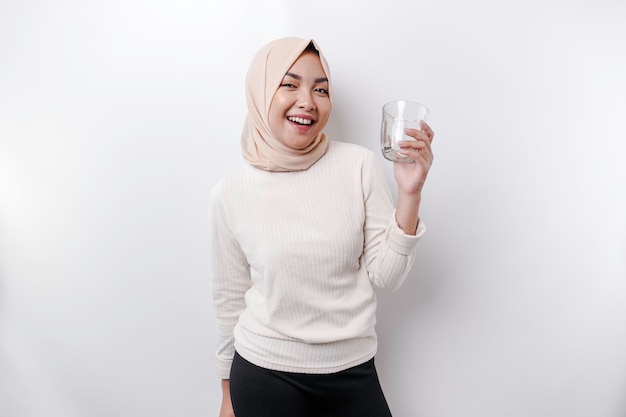 De blije Aziatische Moslimvrouw die hoofddoek draagt drinkt een glas water dat op witte achtergrond wordt geïsoleerd