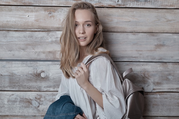 De blonde jonge vrouw van het portret met zilveren rugzak over houten muur