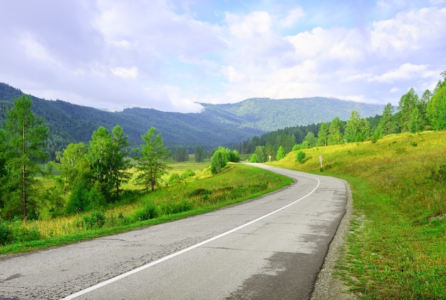 De bocht van de verharde weg tussen de groene bergen met een blauwe bewolkte hemel. Siberië, Rusland